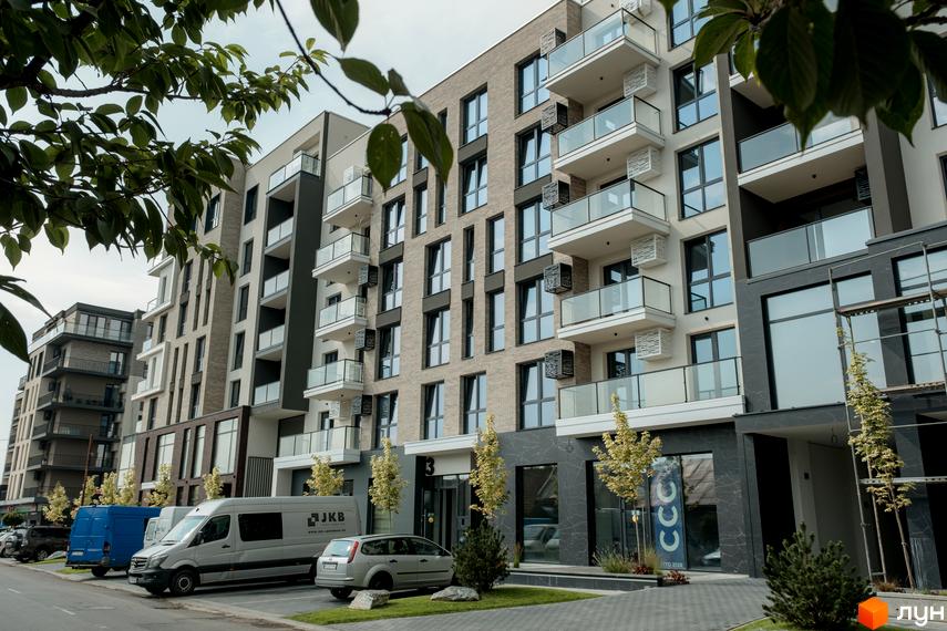 Фасад Житлового комплексу SKY CITY з світлої цегли та темними акцентами, балконами зі скляними поручнями. Перед будинком є зелені насадження та парковка.