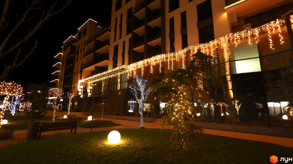 Житловий комплекс SKY CITY з фасадом, прикрашеним святковою ілюмінацією, та доглянутою прибудинковою територією з освітленими деревами та сферичними ліхтарями.
