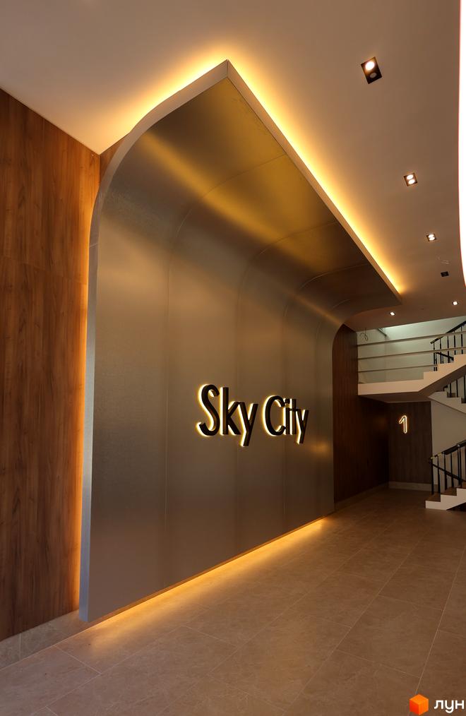 Вхідна група Житлового комплексу SKY CITY: вигнута стіна з підсвіченим логотипом, дерев'яні панелі та світла плитка на підлозі.