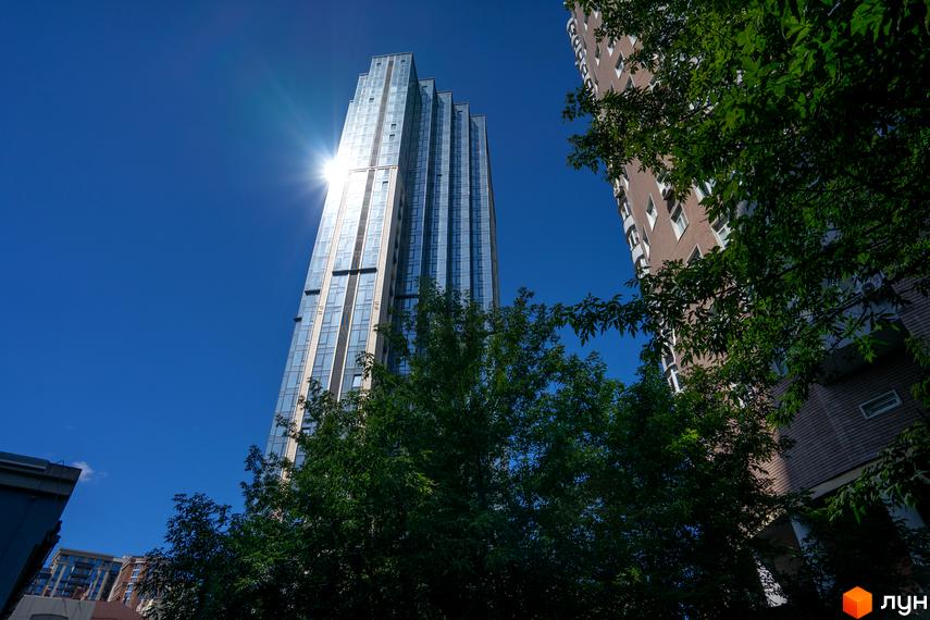 Висотна скляна будівля А136 Highlight Tower з відблиском сонця на фасаді. Поруч розташована цегляна будівля та зелені дерева під яскравим синім небом.