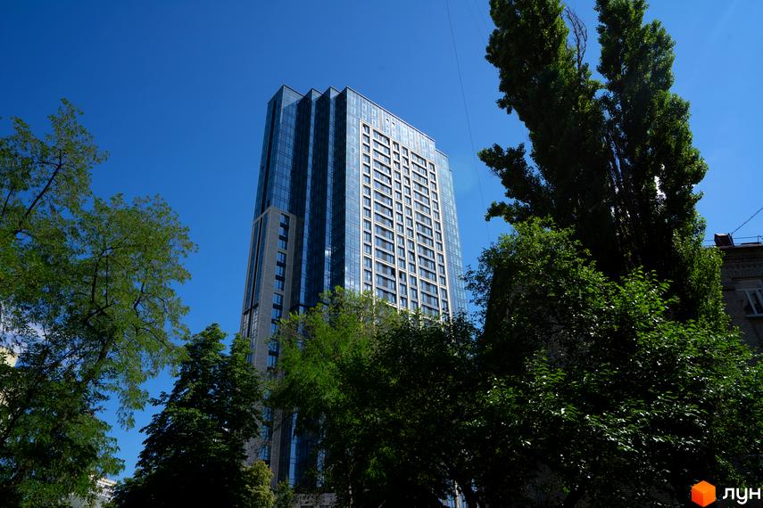 Багатоповерховий будинок А136 Highlight Tower зі скляним фасадом та світлими панелями, оточений зеленими деревами під блакитним небом.