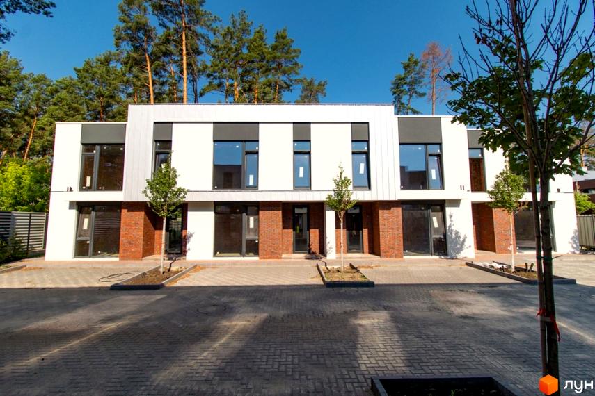 Котеджне містечко КМ At HOME: фасади будинків з цегляними елементами та великими вікнами, перед будинками вимощена парковка та зелені насадження.