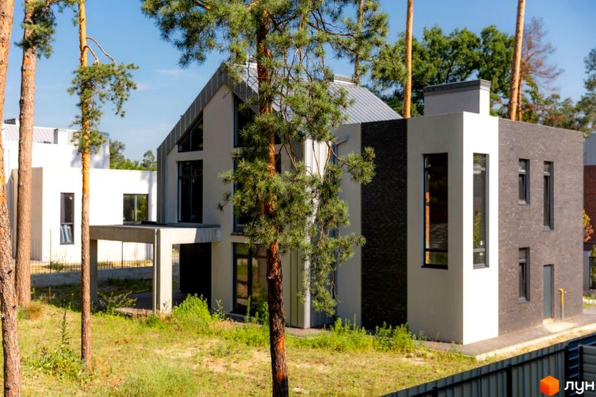 Котеджне містечко At HOME: будинок з білими та темно-сірими фасадами, великими вікнами та сірим дахом. Навколо ростуть високі сосни та зелена трава.