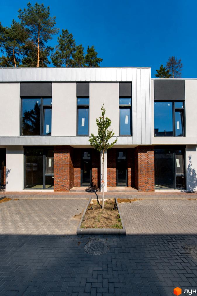 Фасад Котеджного містечка At HOME з світлими стінами, великими вікнами та цегляними вставками. Перед будинками бруківка та зелені насадження.