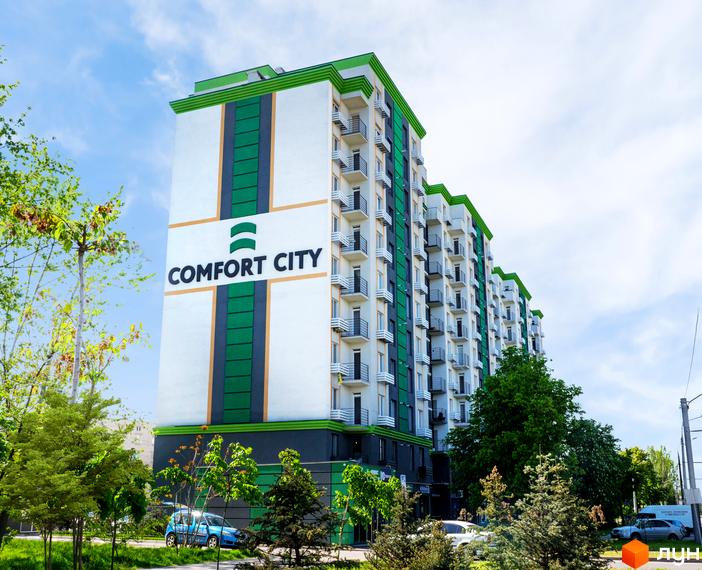 Фасад житлового комплексу Comfort City з зеленими елементами та балконами. Навколо будівлі зелені дерева та автомобіль.