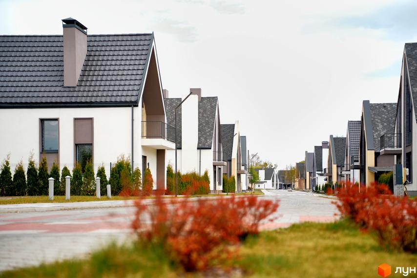 Котеджне містечко КМ BELLAGIO: фасади будинків з темним дахом та білими стінами, огороджені туями та червоними кущами.