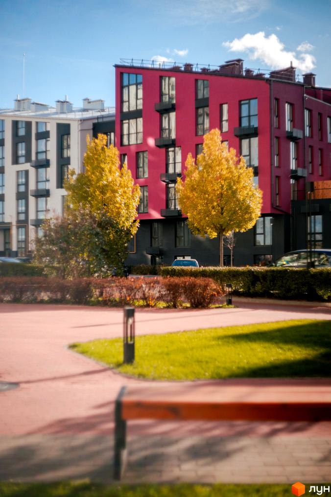 Житловий комплекс City Lake з яскраво-червоною фасадною частиною та сусідніми будинками. Навколо зелені дерева з жовтим листям та доглянута прибудинкова територія з лавкою.