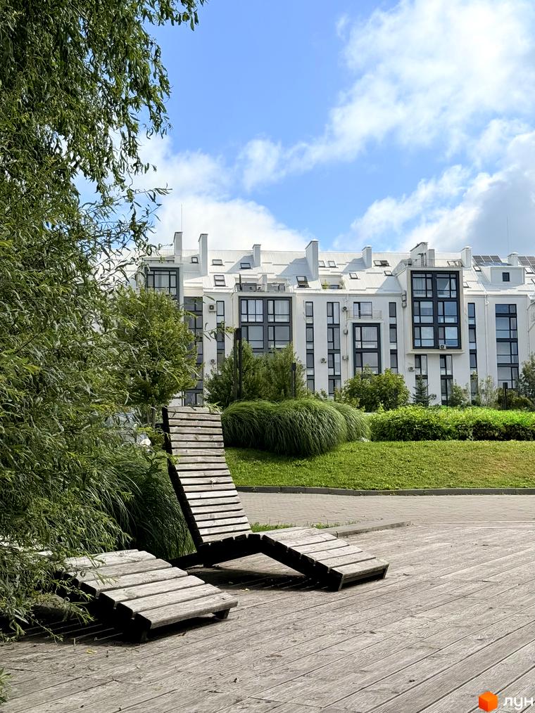 Новобудова City Lake з білим фасадом та великими вікнами, поруч зелена зона з дерев'яними шезлонгами.