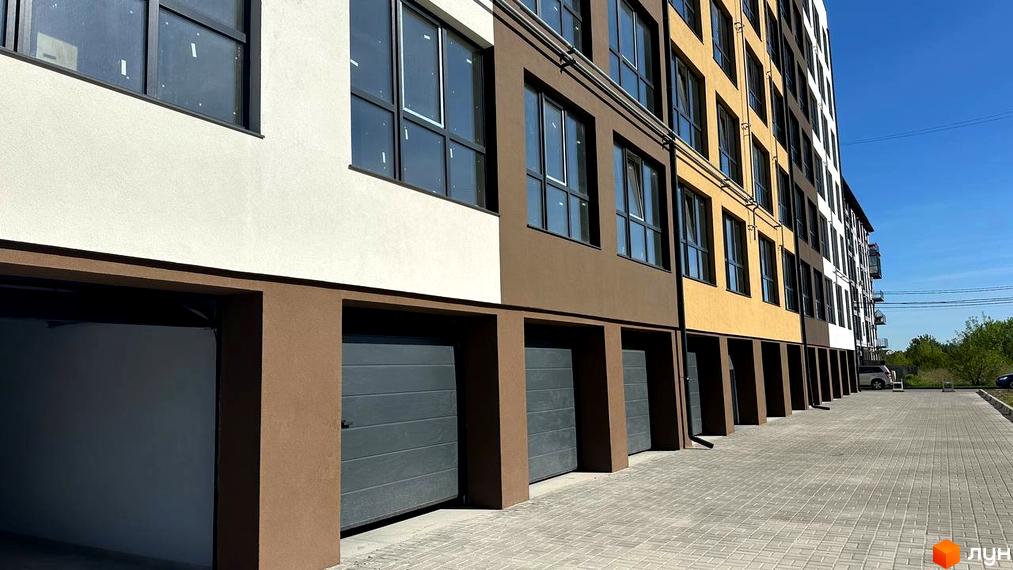 Житловий комплекс PANORAMA DE LUXE: будинок з фасадом у кількох кольорах, гаражами на першому поверсі та вимощеною плиткою територією.