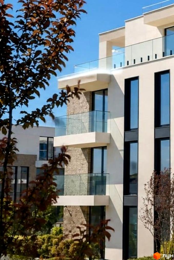 Фасад Житлового комплексу SHERWOOD з бежевими стінами, великими вікнами та скляними балконами. Навколо будинку видно зелені дерева та кущі.