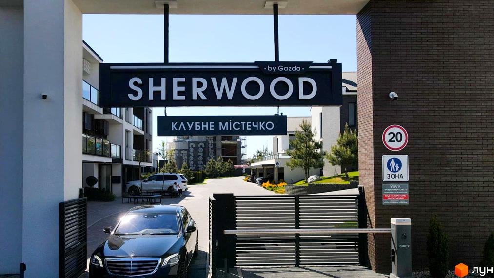 В'їзд до Котеджного містечка SHERWOOD з багатоповерховими будинками світлих відтінків та темними елементами. На території видно доглянутий ландшафт та дорогу.