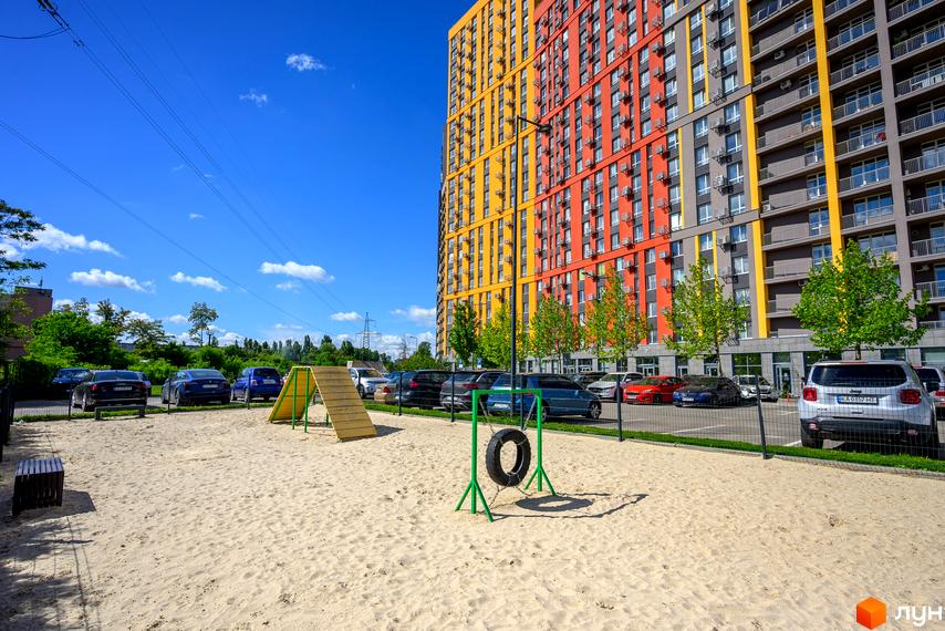 Яскравий багатоповерховий будинок ЖК Svitlo Park та піщаний дитячий майданчик з ігровими елементами на прибудинковій території.