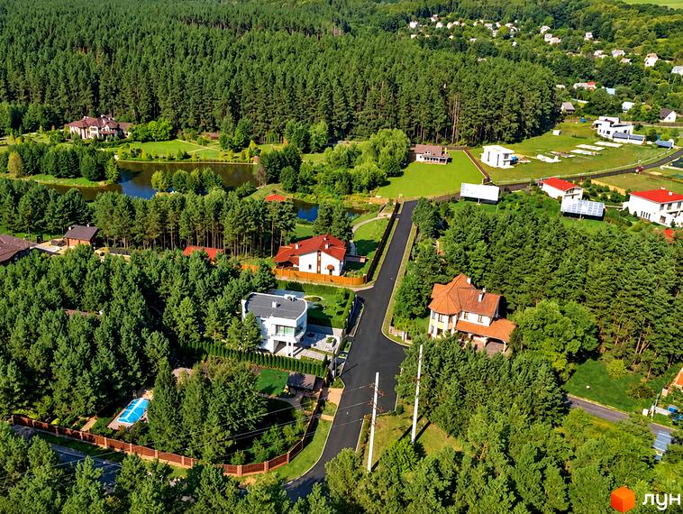 Котеджне містечко КМ Swisstown: двоповерхові будинки з червоними дахами та білими стінами, оточені зеленими деревами та озером. На території є басейн та доглянуті галявини.