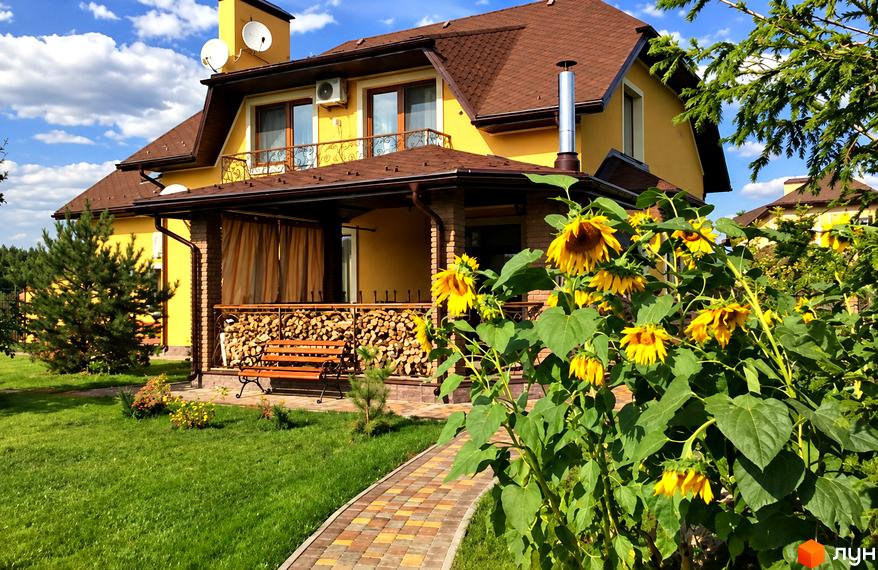 Котеджне містечко КМ Swiss Village: жовтий двоповерховий будинок з коричневим дахом, верандою, дровами та лавкою. Зелений газон, доріжка з плитки та квітучі соняшники.