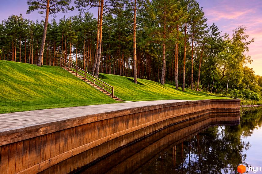 Набережна з дерев'яним пірсом та сходами на зелений схил у КМ Goodlife Park із сосновим лісом вздовж води.
