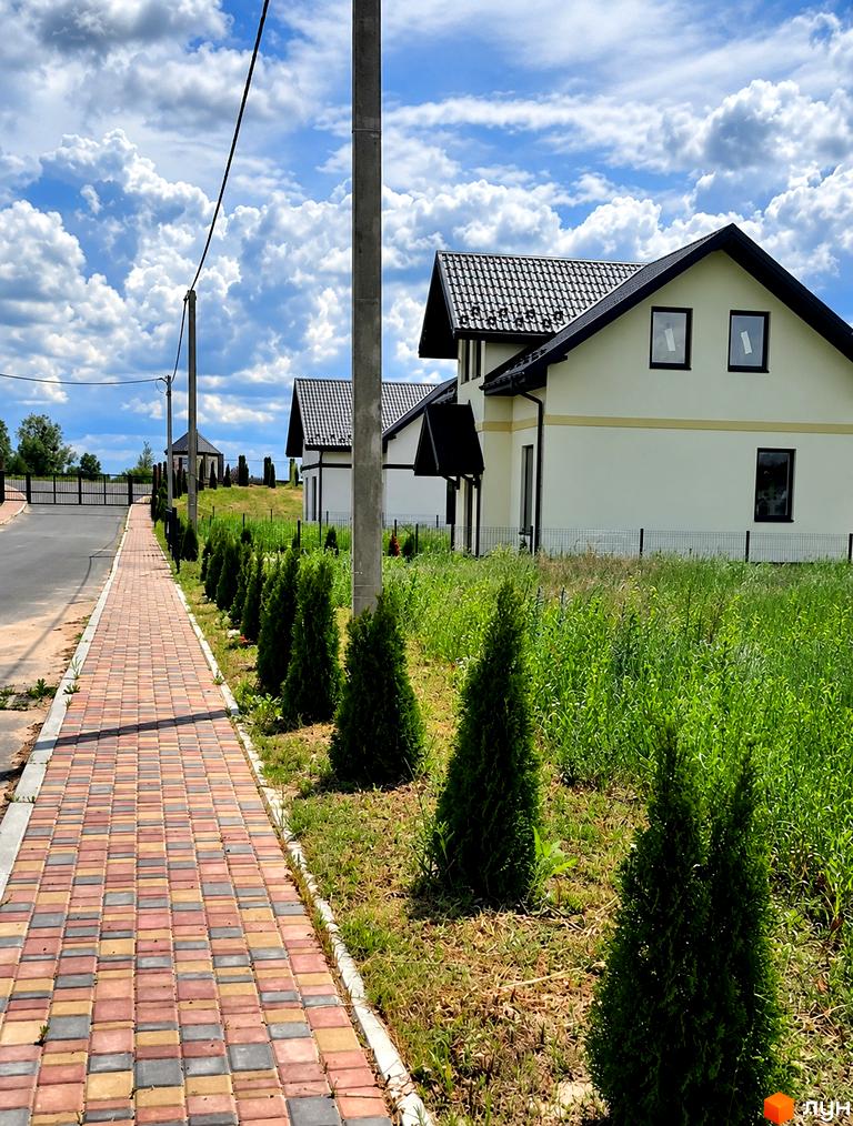 Двоповерховий будинок з білими стінами та чорним дахом у КМ Лелека, біля якого прокладено бруківку та висаджено декоративні туї.