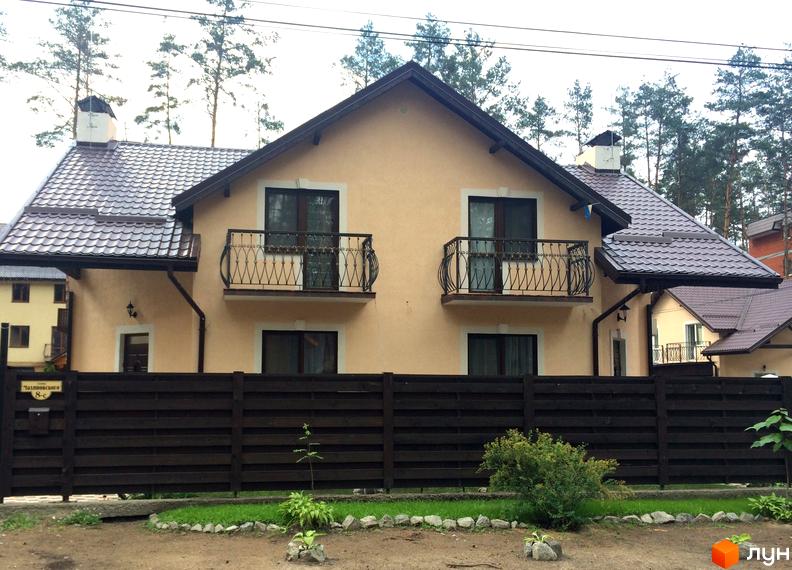 Зовнішній вигляд двоповерхового будинку з балконами та коричневим дахом на вулиці Патона. Прибудинкова територія огороджена темним дерев'яним парканом з елементами ландшафтного дизайну.