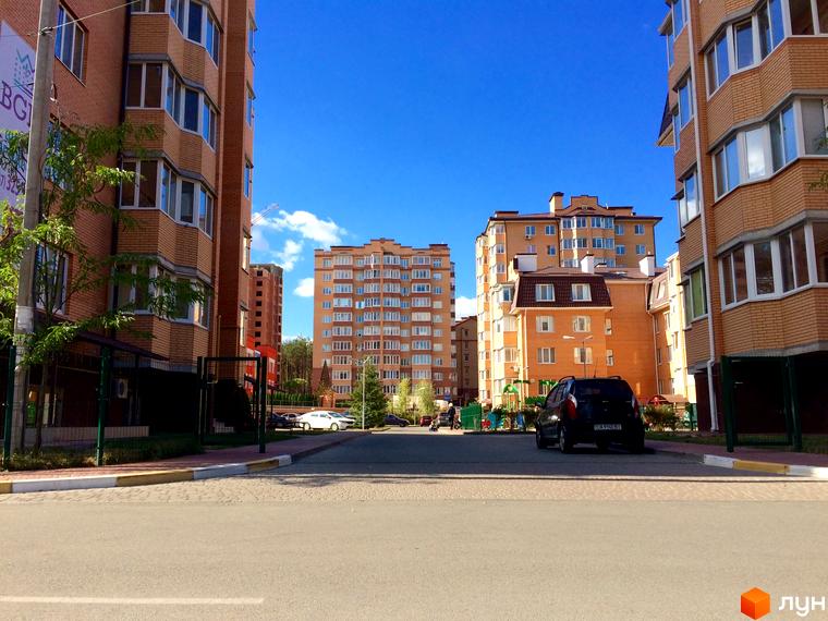 Житловий комплекс Південний - це цегляні будинки з еркерними вікнами під блакитним небом. На прибудинковій території є дитячий майданчик та автомобілі.