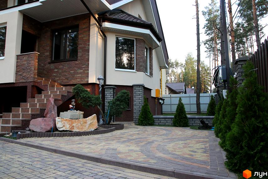 Будинок з цегляним оздобленням та сходами, що ведуть до входу. Прибудинкова територія з бруківкою та зеленими насадженнями.