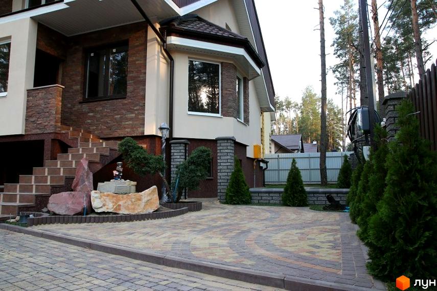 Фасад будинку з цегляним оздобленням та сходами, прибудинкова територія з бруківкою та зеленими насадженнями.
