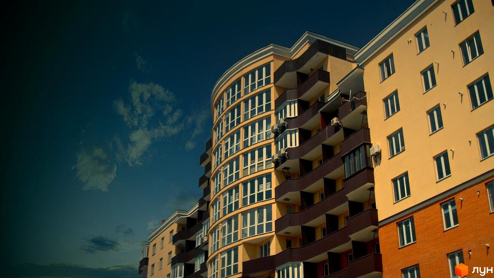 Новобудова на вулиці Івана Мазепи, 11. Будинок має жовтий фасад, вигнуту форму та коричневі балкони. Навколо будинку видно блакитне небо з хмарами.