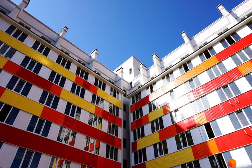 Житловий комплекс Олексіївські Акварелі з яскравим фасадом з жовтими, помаранчевими та червоними елементами. Багатоквартирний будинок з великою кількістю вікон під блакитним небом.