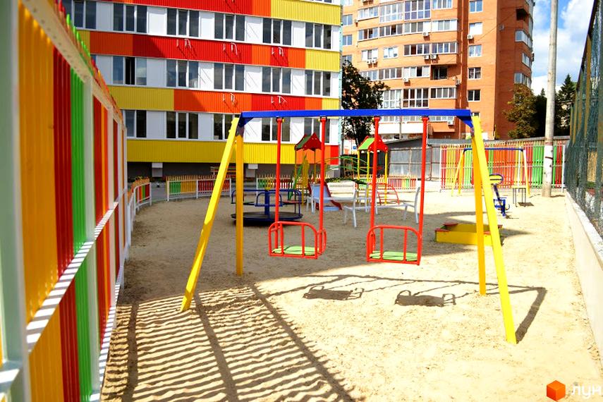 Житловий комплекс Олексіївські Акварелі: яскравий фасад будинку та дитячий майданчик з гойдалками та пісочницею.
