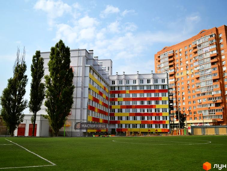 Житловий комплекс Олексіївські Акварелі: яскрава фасадна частина будівлі з червоними, жовтими та білими смугами, розташована поруч з футбольним полем та зеленими деревами.