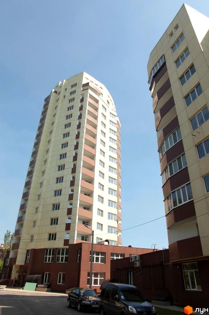 Житловий комплекс Олександрівський посад - високі світлі новобудови з коричневими елементами та балконами. Навколо будівельний майданчик з автомобілями.