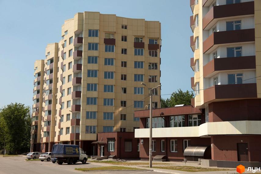 Житловий комплекс Олександрівський посад - висотні будинки світло-бежевого кольору з коричневими елементами та балконами. На прибудинковій території видно автомобілі та вантажівку.