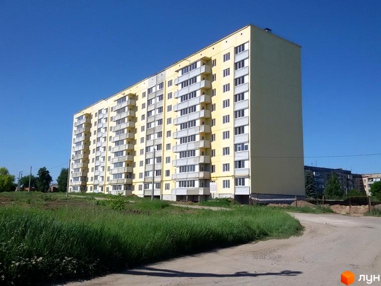 Зовнішній вигляд жовтої багатоповерхівки з балконами на вулиці Нафтовиків, 15/1. Навколо будинку зелена трава та ґрунтова дорога.