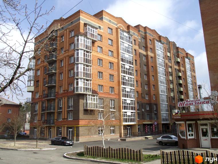 Багатоповерховий цегляний житловий комплекс з еркерними балконами та великими вікнами. Поруч розташована прибудинкова територія з деревами та парканом.