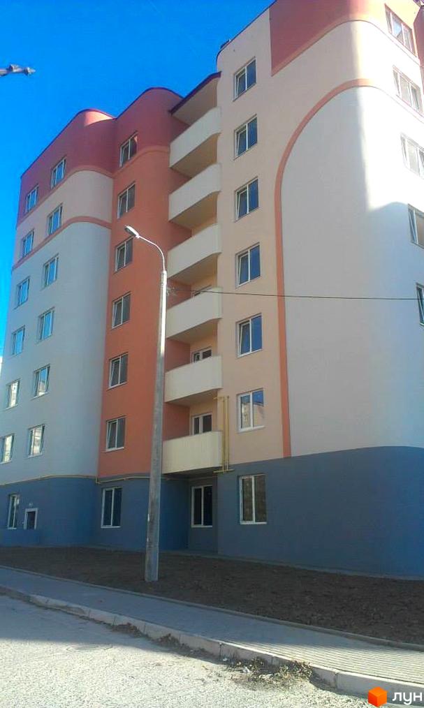 Новобудова Родинний: багатоповерховий будинок з бежевими, теракотовими та синіми фасадами, закругленими кутами та балконами. Поруч тротуар та ґрунт.