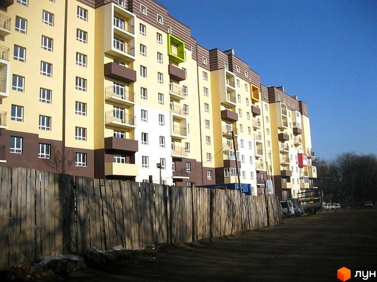 Житловий комплекс Родинний затишок: багатоповерховий будинок світло-жовтого та коричневого кольорів з балконами. Перед ним дерев'яний паркан та ґрунтова дорога.