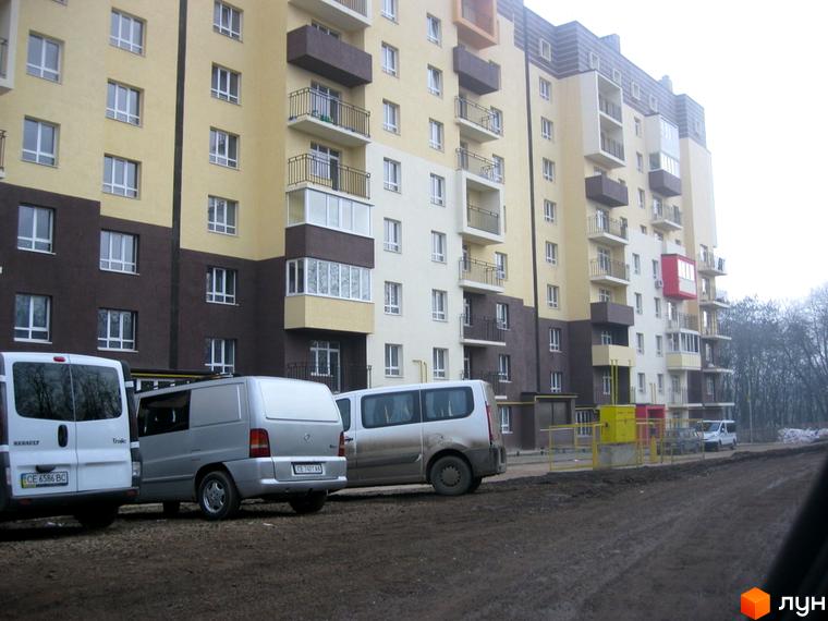 Житловий комплекс Родинний затишок: багатоповерховий будинок з бежево-коричневим фасадом та балконами. На передньому плані ґрунтова дорога та автомобілі.