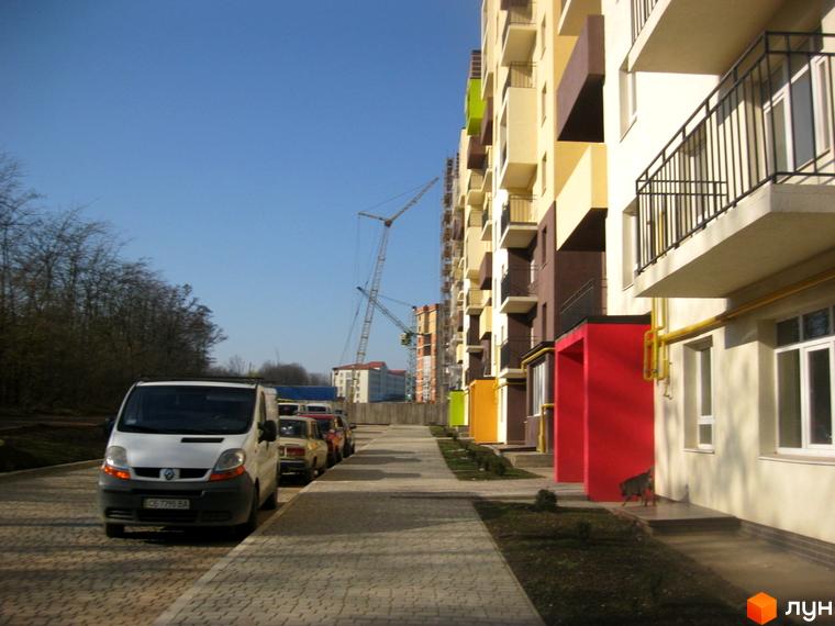 Багатоповерховий будинок Житлового комплексу Родинний затишок з бежевим фасадом та кольоровими акцентами. Прибудинкова територія з доріжкою та припаркованими авто.