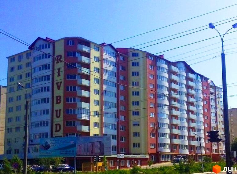 Житловий комплекс Rivbud: багатоповерховий будинок з червоно-жовтими фасадами та білими балконами. Перед ним зелена зона з дорогою та автомобілями.