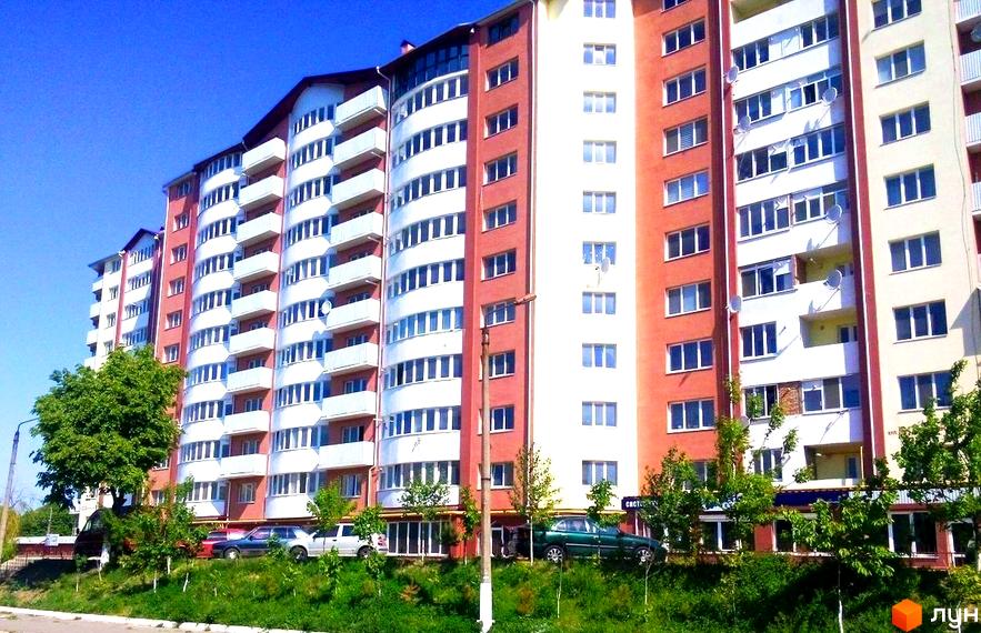 Житловий комплекс Rivbud: багатоповерховий будинок з червоно-білим фасадом та балконами. Прибудинкова територія з зеленою травою та деревами.