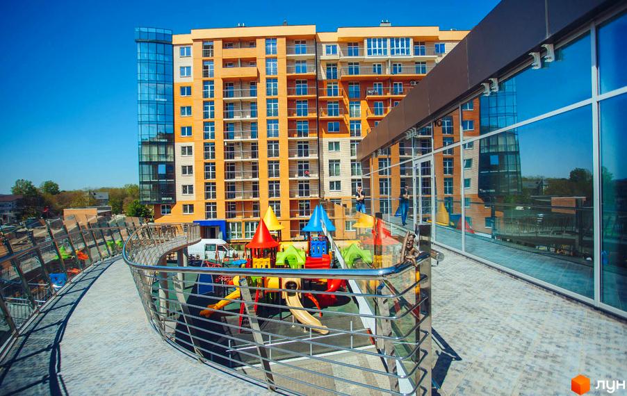 Житловий комплекс Водограй: багатоповерховий будинок з помаранчевими фасадами та скляними елементами. На території яскравий дитячий майданчик та пішохідний міст.