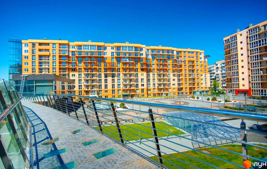 Багатоповерхові будинки Житлового комплексу Водограй з помаранчевими фасадами та балконами. Прибудинкова територія має пішохідний міст, зелені газони та зони відпочинку.