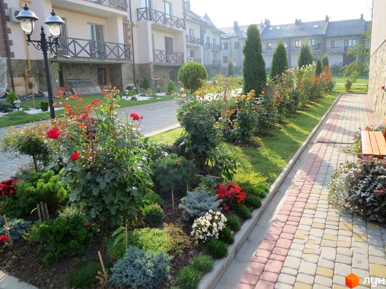 Котеджний комплекс КМ Віденська брама з доглянутою прибудинковою територією та квітучими рослинами. Фасади будівель виконані в світлих тонах з дерев'яними елементами.