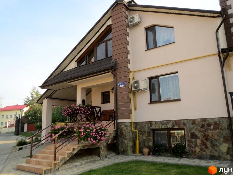 Будинок Котеджного містечка Віденська брама з бежевим фасадом, коричневим дахом, кам'яним цоколем та великим ґанком зі сходами. Прибудинкова територія має зелений газон та квіти.