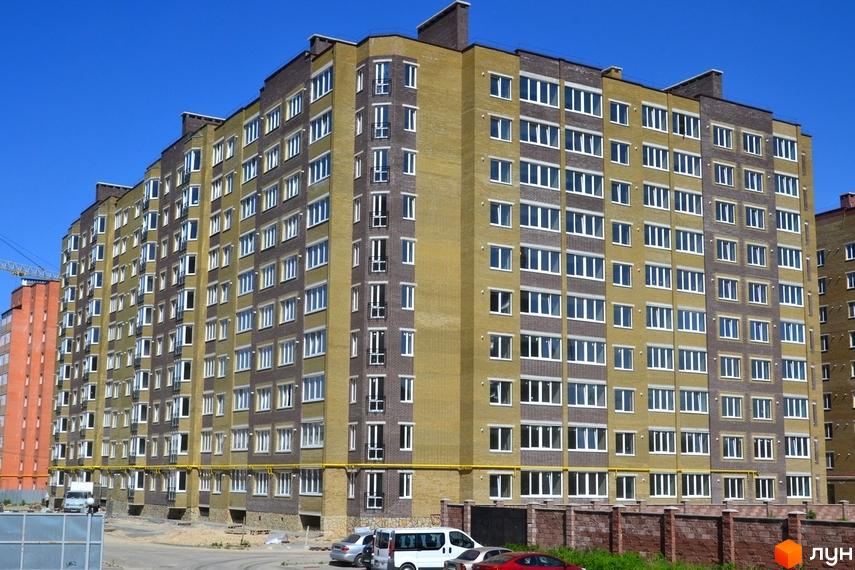 Фасад новобудови на вулиці Іващенка, 1а, з цегляним оздобленням та великою кількістю вікон. Поруч видно прибудинкову територію з автомобілями та огорожею.