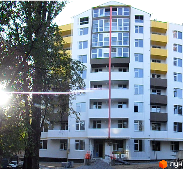 Фасад новобудови ЖК Фенікс з жовтими балконами та численними вікнами. Навколо будівлі видно дерева та будівельні роботи.