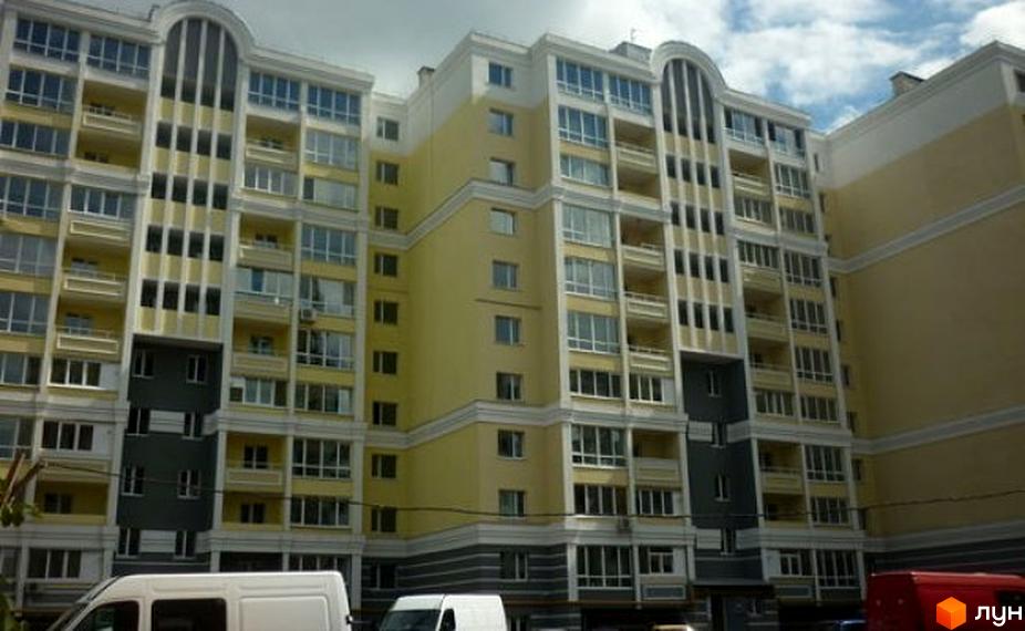 Фасад багатоповерхового будинку в жовтих та сірих тонах, з численними вікнами та балконами. На передньому плані видно білий фургон.