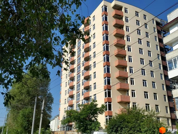 Новобудова на вулиці Сахарова. Багатоповерховий будинок світло-бежевого кольору з цегляними балконами та великими вікнами. Навколо будинку зелені дерева та кабелі.