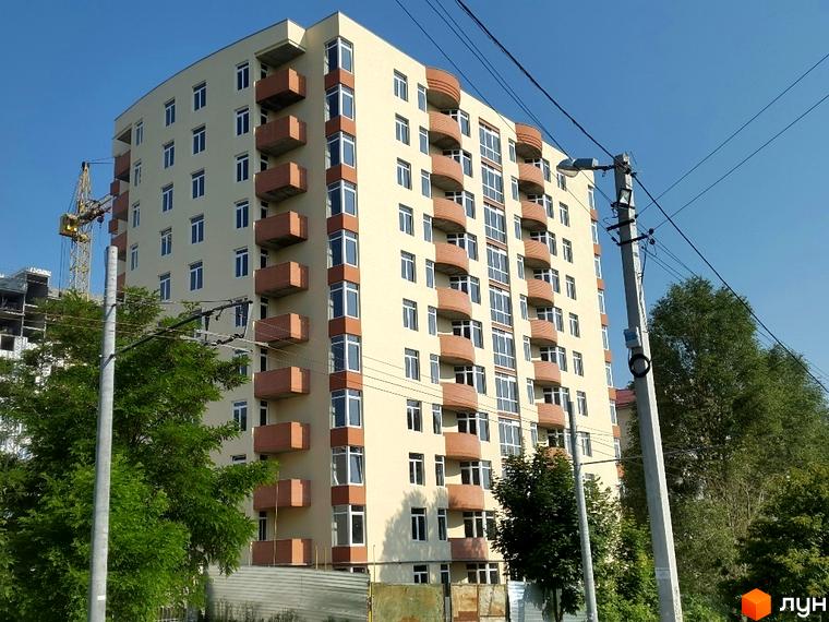 Будівельний проєкт на вулиці Сахарова. Багатоповерховий будинок світло-бежевого кольору з коричневими балконами та великими вікнами. Навколо зелені дерева та елементи міської інфраструктури.