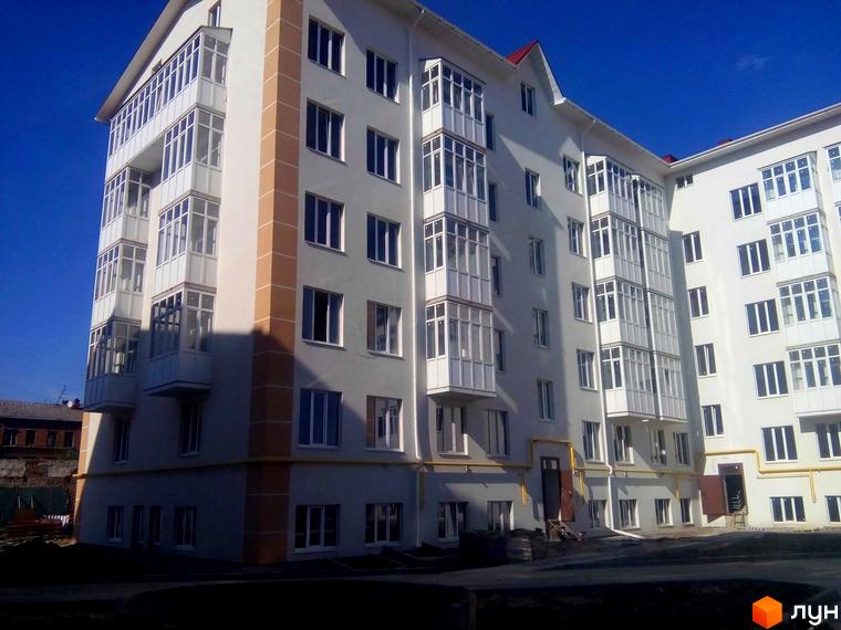 Зовнішній вигляд житлового комплексу на вул. Дмитрівська, 19. Білий багатоповерховий будинок з численними заскленими балконами та коричневими вертикальними смугами. Навколо будинку видно будівельні роботи.