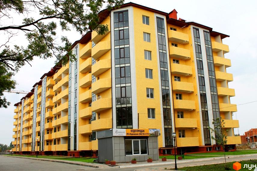 Високий будинок з жовтими стінами та балконами. Поруч розташована прибудинкова територія з асфальтованою дорогою та зеленими насадженнями. ЖК Маріїнський.