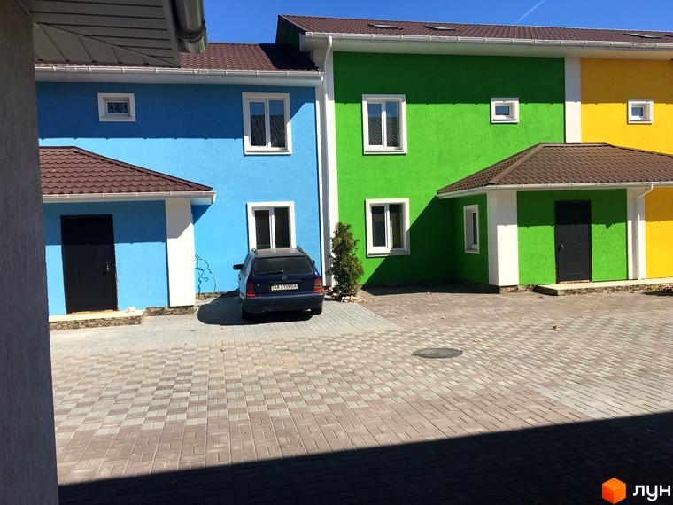 Комплекс Ivankiv-townhouse: фасади будинків у блакитному, зеленому та жовтому кольорах. Прибудинкова територія з бруківкою та автомобілем.